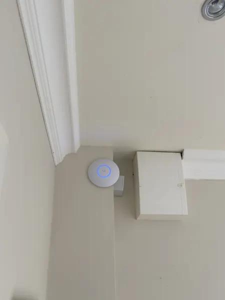 White round Wi-Fi access point with a blue light ring fixed high on a wall at the ceiling line, beside a cream coloured junction box