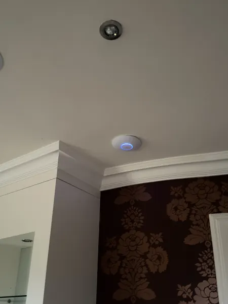White round Wi-Fi access point with a blue light ring fixed to a ceiling beside a recessed light, above brown patterned wallpaper