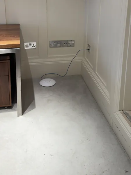 White round Wi-Fi access point lying on the floor beside a desk, with a thin blue cable running up to a wall outlet