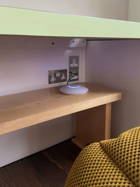 White round Wi-Fi access point with a blue light ring on a wooden shelf beside a wall socket, with a green cable running to a wall outlet