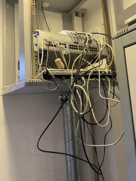 Wall mounted equipment frame with network switches and a patch panel, and loose white, black and blue cables hanging down below it