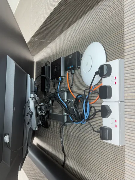 Recess behind a wall mounted TV with two double sockets, a white Wi-Fi access point, a Blustream unit, power supplies and orange and blue network cables