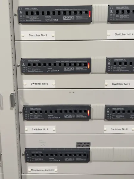 Close up of Philips Dynalite 12 channel relay controllers labelled Switcher No.3 to No.8, with a handwritten label above the miscellaneous controller reading gym voile open and close