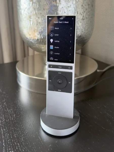 Handheld remote with a colour screen listing watch, listen, lighting, shades and comfort for a guest bedroom, standing on its charging base beside a silver glass lamp