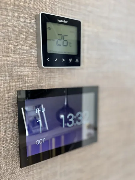 Heatmiser thermostat showing 26 degrees above a black wall touch screen that shows the date and time