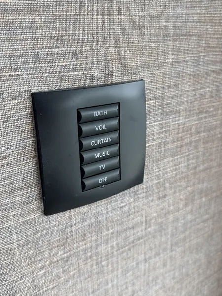Black wall keypad with six buttons labelled bath, voil, curtain, music, TV and off, on a grey woven wall covering