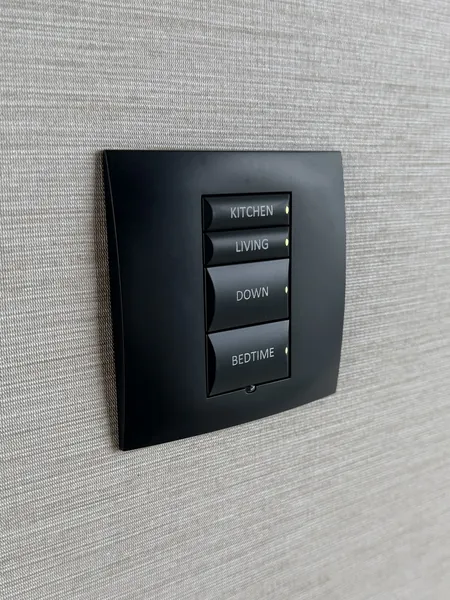 Black wall keypad with four backlit buttons labelled kitchen, living, down and bedtime, on a pale grey wall covering