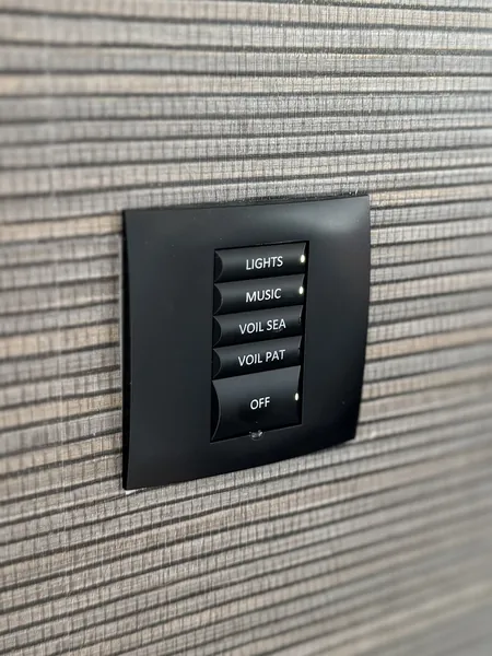 Black wall keypad with buttons labelled lights, music, voil sea, voil pat and off, on a woven wall covering