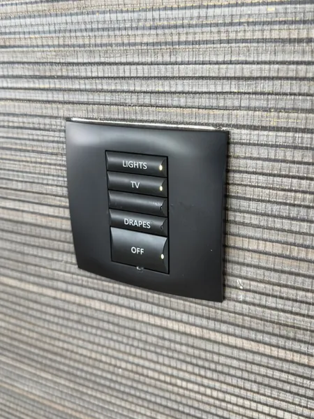 Black wall keypad on a dark woven wall covering, with buttons labelled lights, TV, drapes and off and one blank button