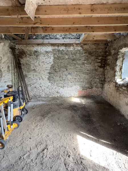 The cottage stripped back to a bare stone shell with new ceiling joists overhead and tools on the earth floor