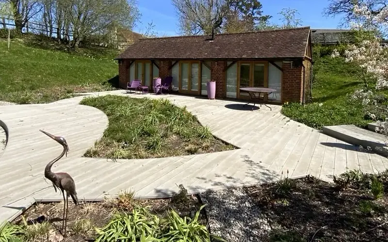 Single storey brick garden building with glazed doors, beside a curved pale timber deck and a heron sculpture