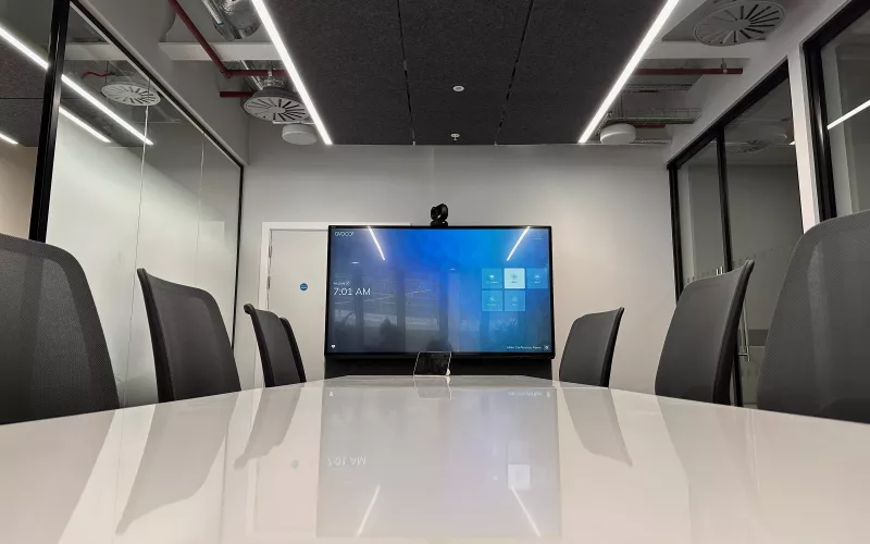 View along a glossy white table to a screen and camera in a glass walled meeting room
