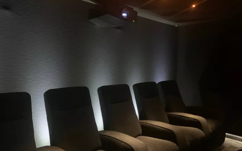 Home cinema lighting at work: a row of dark recliners in front of a textured wall bathed in white light, with the projector mounted near the ceiling at the back