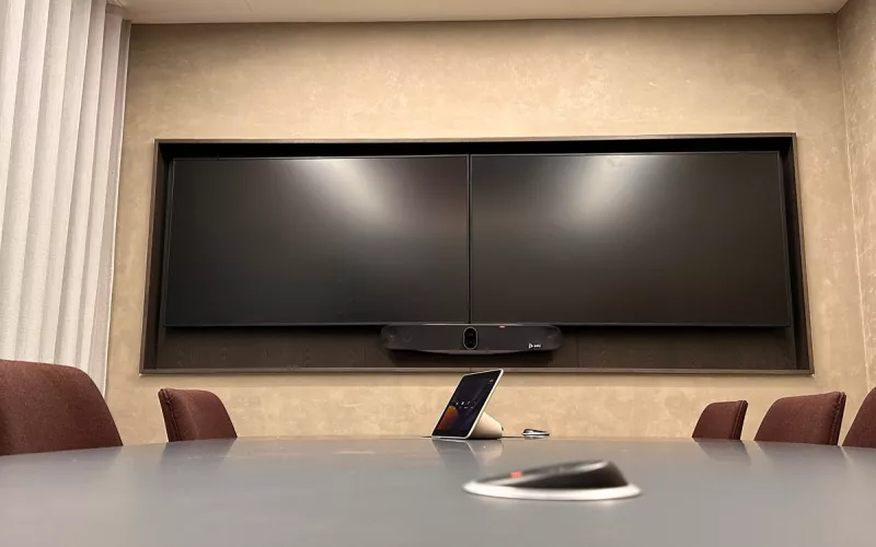A Poly video bar below two displays set side by side in a wall recess, with a touch controller on the meeting table