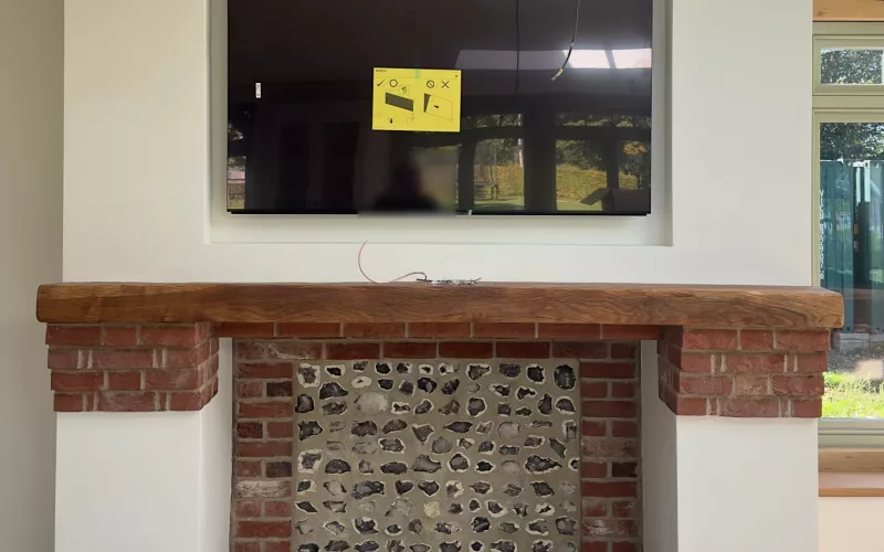 Recessed TV installation above an oak beam mantel, with the screen sitting into the wall over a brick and flint fireplace