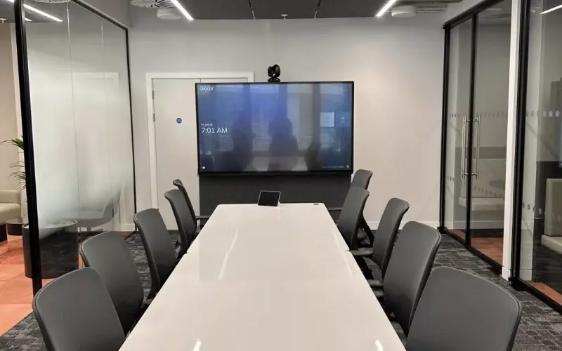 Square view of a meeting room behind glass, showing acoustic ceiling panels and linear lights above a white table, dark chairs, a touch panel and a display on the wall with a camera over it. Rooms like this are where a Teams Rooms Android audit starts