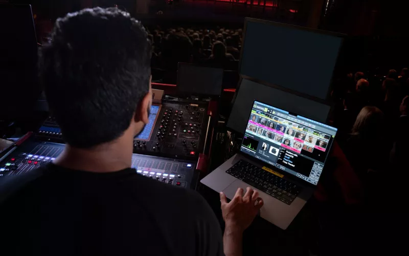 Front of house mixer in a theatre with a laptop showing Shure WAVETOOL 4 channel tiles and a chat message, audience in the background