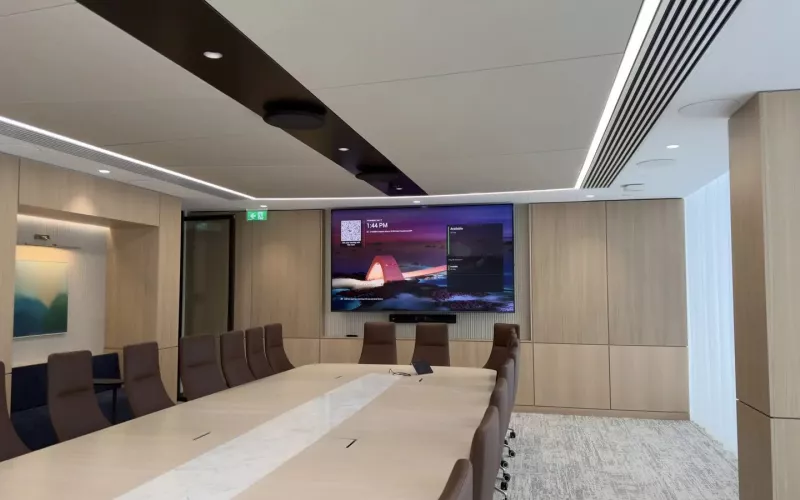 Wide view of a boardroom: a long pale table with a marble inlay, brown leather chairs, a wall mounted display, microphones fixed to the ceiling and a linear light. Larger rooms like this are where the Biamp Devio UCX 2000 and 3000 are aimed