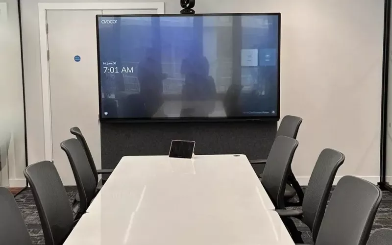 Wide view of a meeting room: a display on the wall with a camera mounted over it, a long white table with a small touch panel and dark chairs, a typical setting for Neat Teams certified room controllers