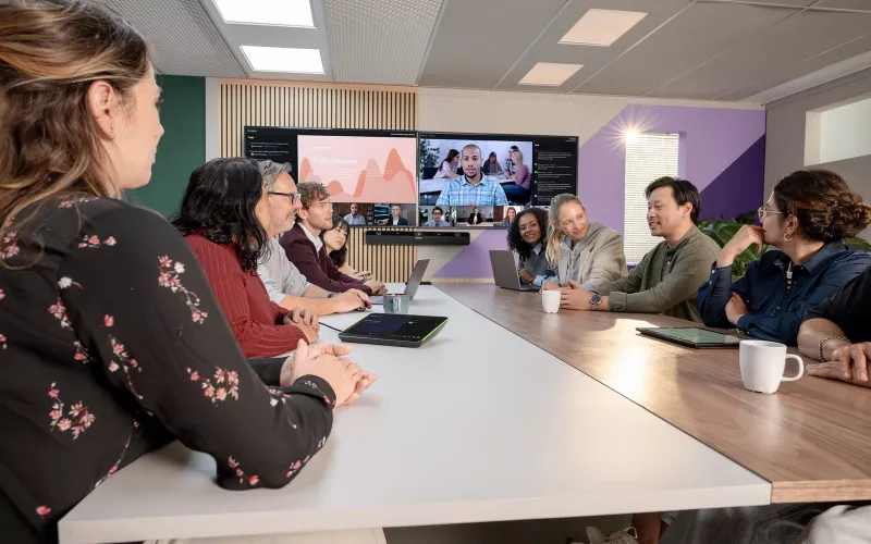 Meeting room with a team around a table and a Shure IntelliMix Bar Pro mounted below two wall displays, with the touch panel on the table, as used in Dublin and Cork Teams Rooms