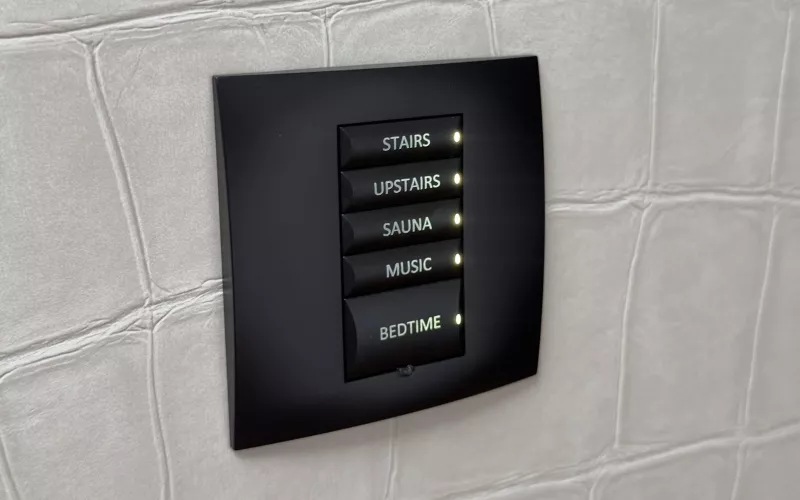 Wall keypad in a Cork house, part of the Control4 to Crestron Home project, with backlit buttons labelled stairs, upstairs, sauna, music and bedtime on a pale crocodile pattern wall