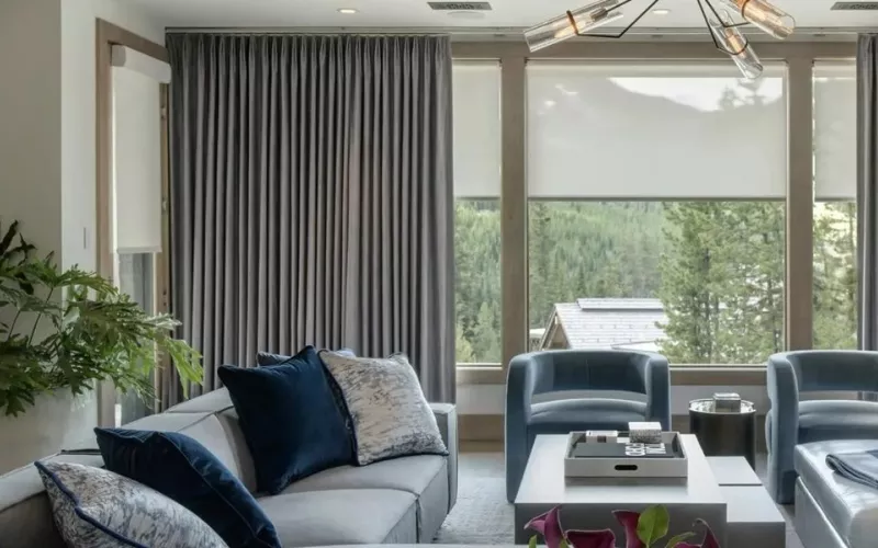 Living room with floor length grey curtains and pale roller shades lowered part way over a wide window, the layered look that Crestron shades can automate