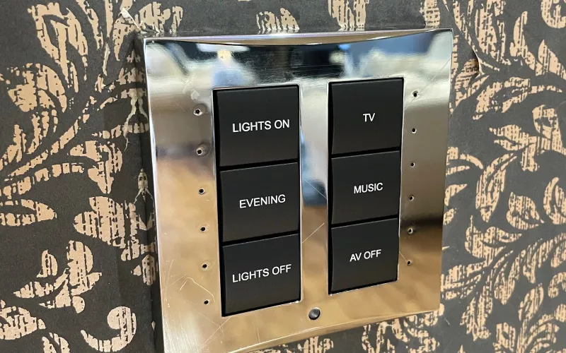 Polished chrome keypad from a home automation system on damask wallpaper, with engraved buttons for lights on, evening, lights off, TV, music and AV off