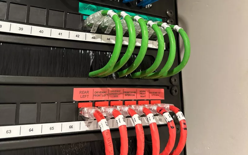 Security wiring in a home network rack: red patch leads for the cameras and the front door intercom, each labelled by location, below a row of green data leads