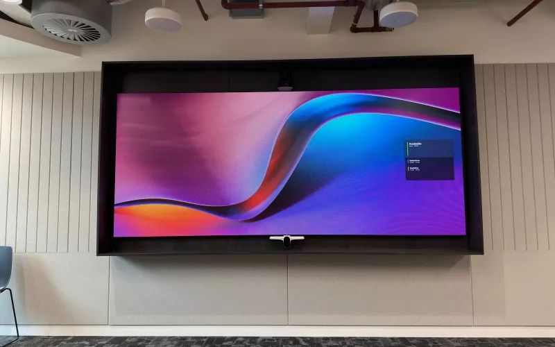 Large screen in a dark lined recess with a camera above, a video bar below and two white pendant speakers overhead, the kind of equipment meeting room AI depends on