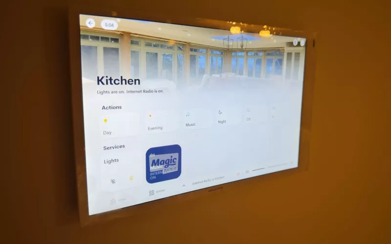 Wall mounted Crestron touch screen showing a kitchen page with day, evening, music and night scenes, the kind of screen that can also show heat pump controls