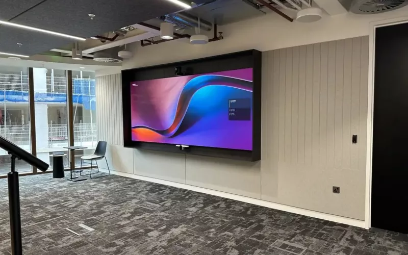 Large wall screen with a camera above and a bar below in a dark surround, in an open plan office with a stair rail, the kind of wall where interactive displays are fitted