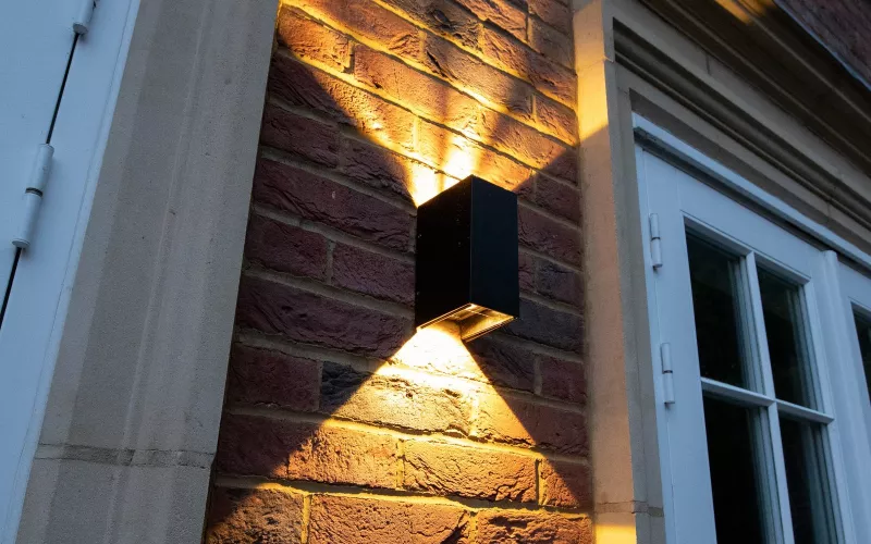 Black up and down wall light washing warm light over a brick wall between two stone framed doors at dusk, the kind of front wall where EV charger wiring is planned