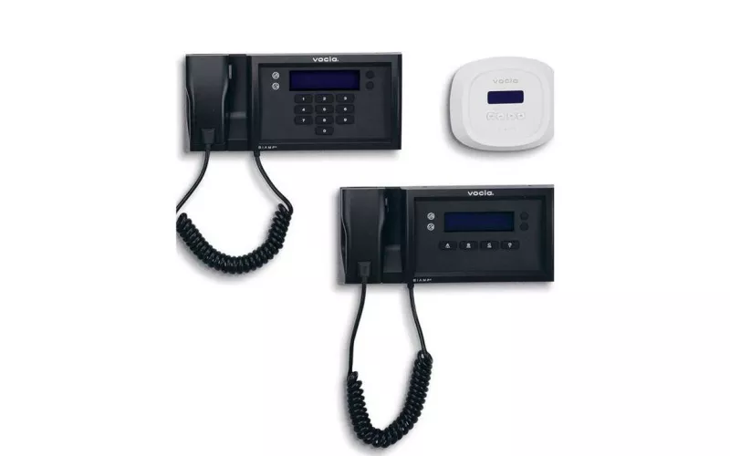 Biamp Vocia wall mounted paging stations in black with coiled handsets and backlit displays, one with a number keypad, beside a small white Biamp Vocia wall controller