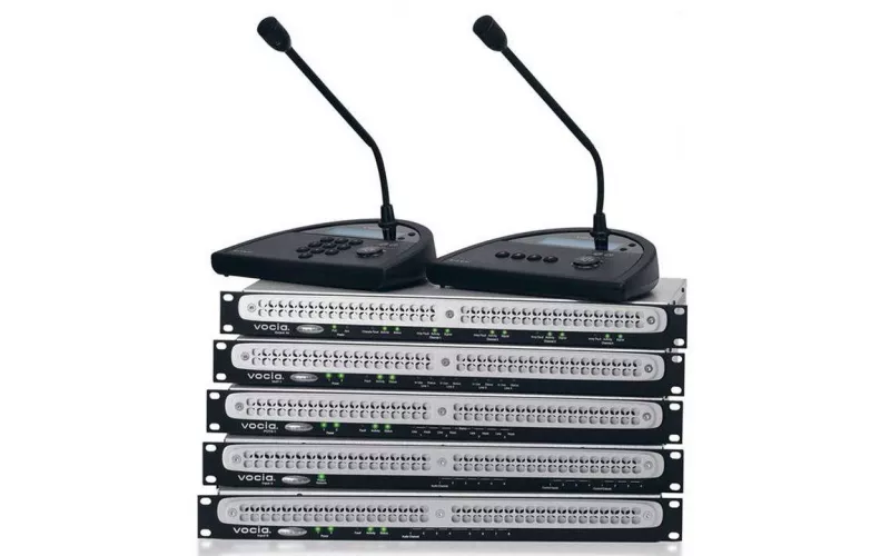 Two Biamp Vocia desk paging stations with gooseneck microphones, one with a number keypad, sitting on a stack of five rack mounted Biamp Vocia units