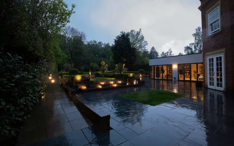 Wet stone terrace with lit garden steps at dusk beside a flat roofed garden wing, the kind of outbuilding roof that can carry solar panels on a larger site