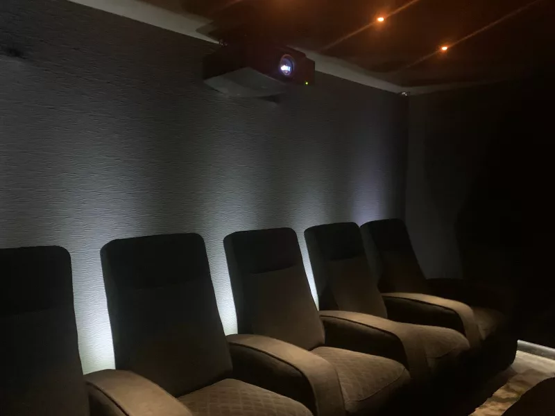 Home cinema lighting at work: a row of dark recliners in front of a textured wall bathed in white light, with the projector mounted near the ceiling at the back
