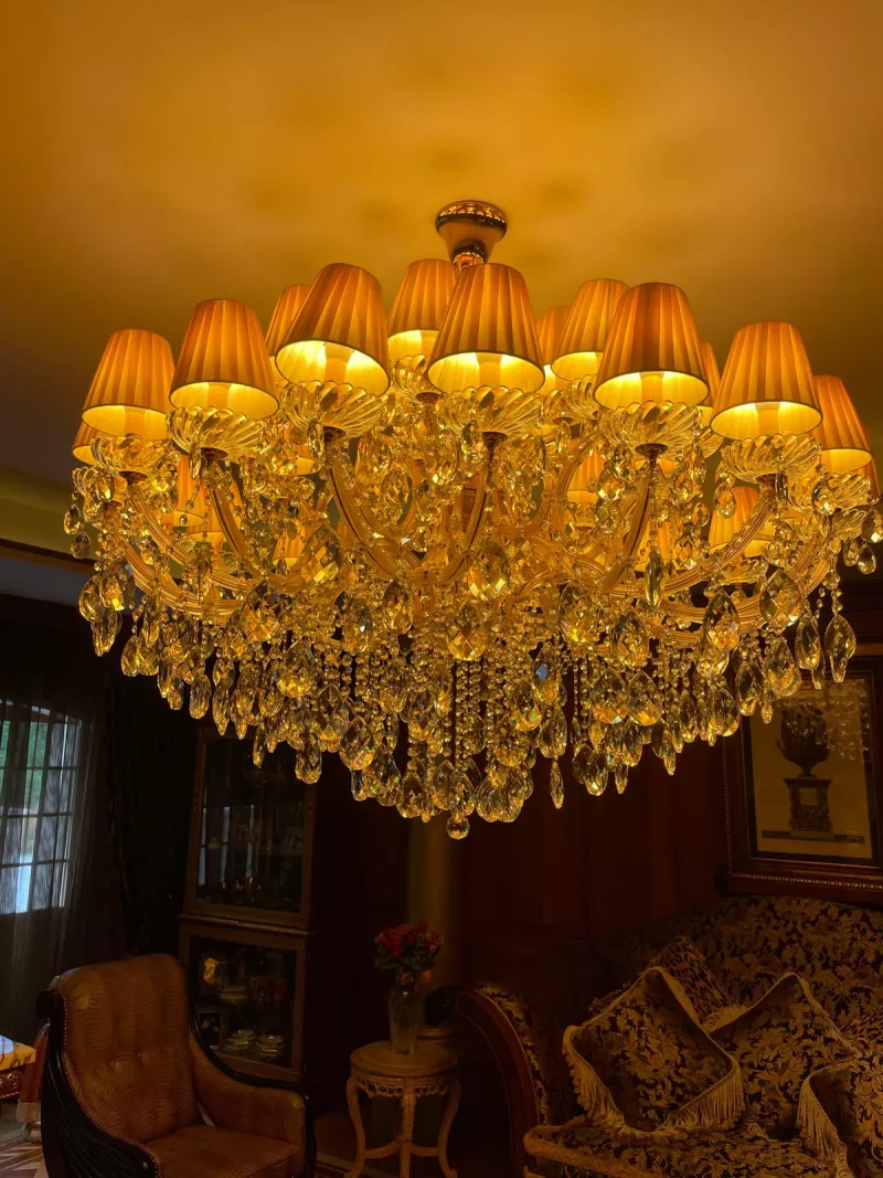 Pleated cream shades and hanging crystals on a chandelier above a sitting room of dark furniture and patterned sofas