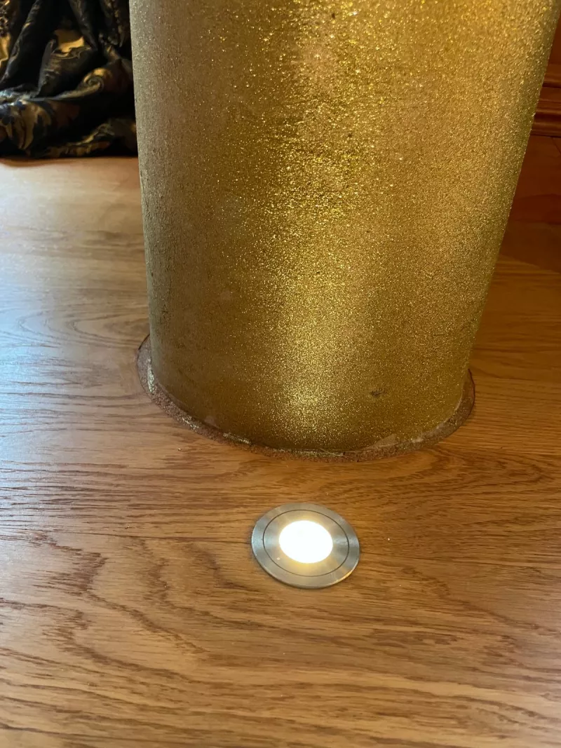 A small round LED fitted into oak floorboards at the foot of a gold column