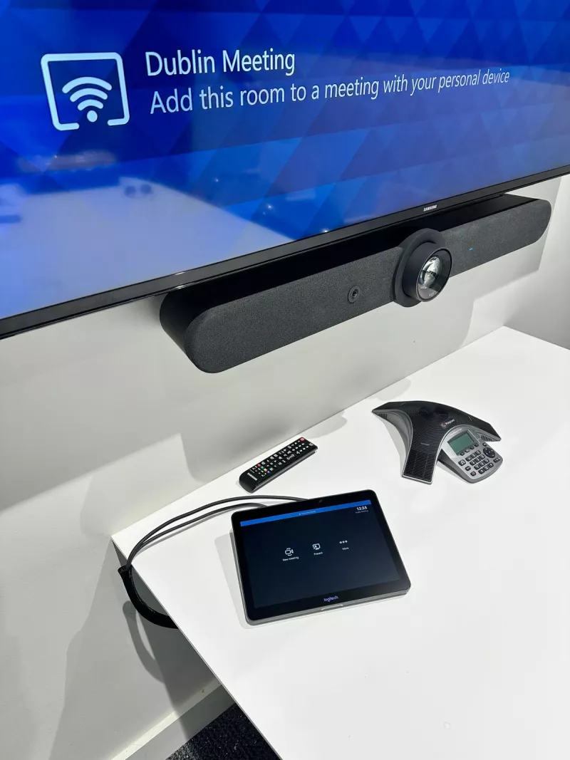 Close view of the Logitech video bar below the display, with a Logitech Tap touch controller and a speakerphone on the table
