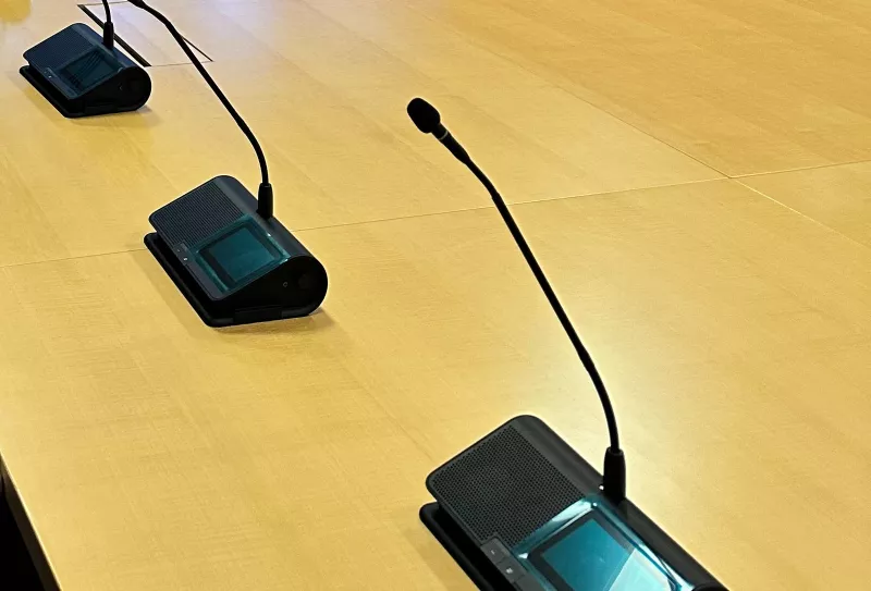 Three conference microphone units on the timber table, each with a gooseneck microphone, a small screen with buttons and a speaker grille