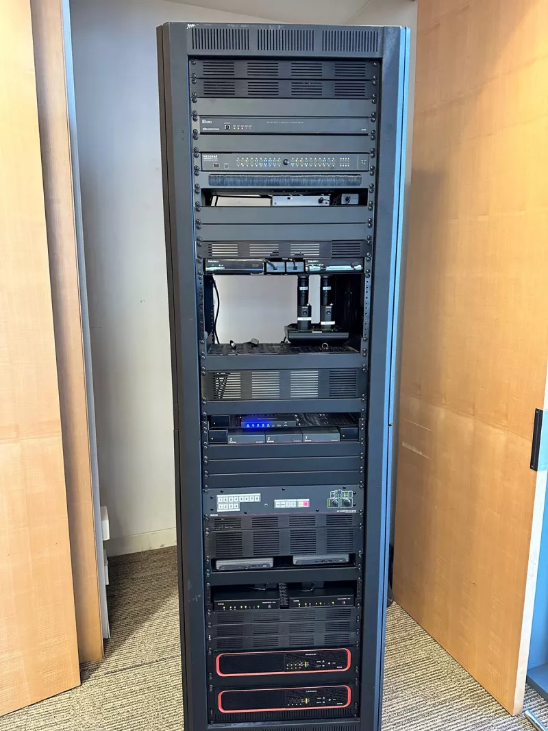 The full equipment rack in its cupboard, from the Crestron control processor at the top to the two Biamp amplifiers at the bottom, with Shure wireless microphones on a shelf in the middle