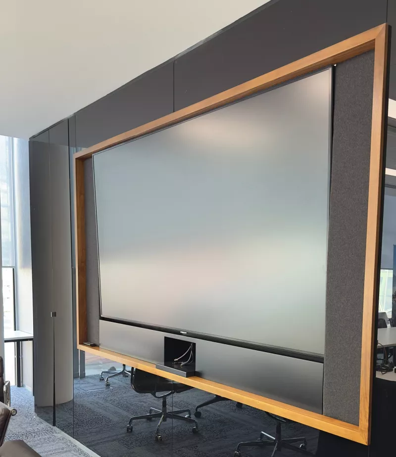 A large screen set into a timber frame with grey fabric panels on each side, on a black glass wall
