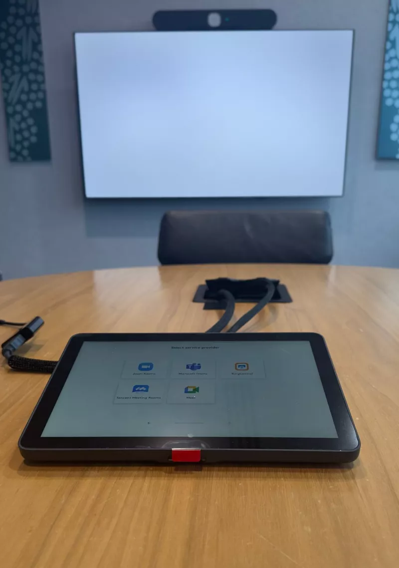 A Logitech Tap touch controller on a round timber table, asking which meeting service the room should use, with the screen and video bar behind