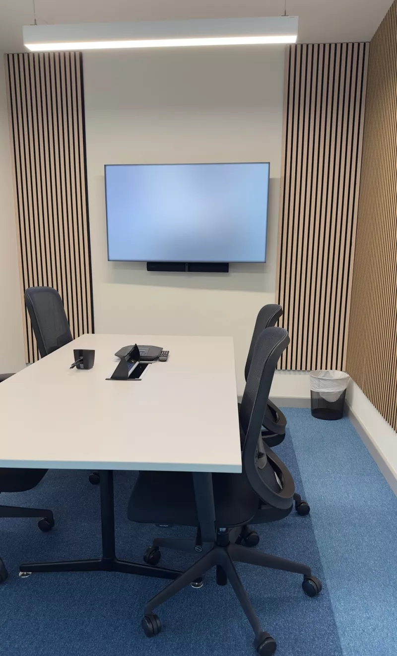 A small meeting room with timber slat acoustic panels either side of the screen, a slim video bar below it and a conference phone on the white table