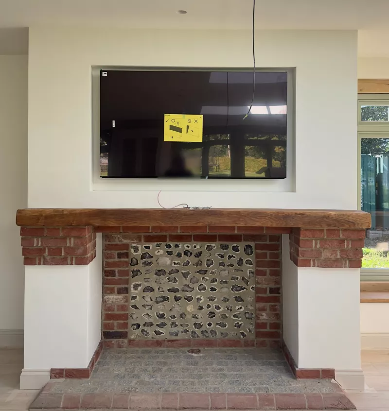 Recessed TV installation above an oak beam mantel, with the screen sitting into the wall over a brick and flint fireplace