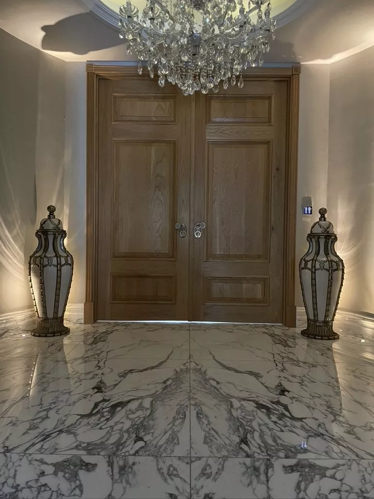 Wall touch panel beside oak double doors, under a crystal chandelier in a marble floored hall