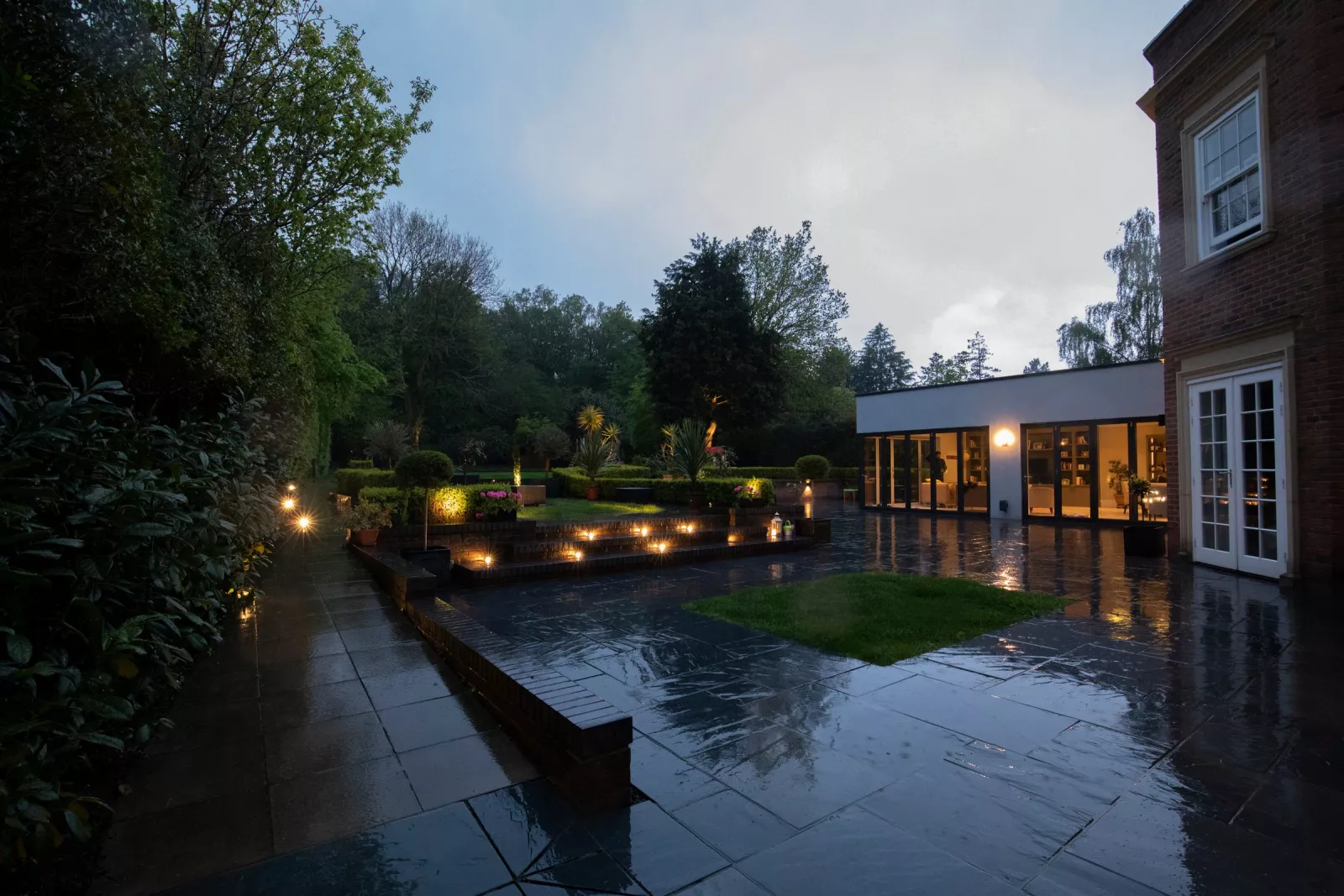 Paved garden terrace and lawn lit at dusk in front of a lit glass extension, the kind of house and garden a KNX installer in Ireland puts on one system
