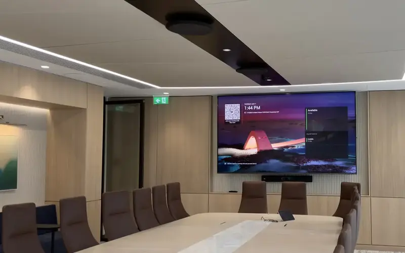 Meeting room with a wide screen on the end wall and a long pale table under recessed lighting