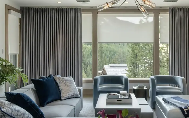 Sitting room with tall grey curtains beside a large window, two blue armchairs and a low white table