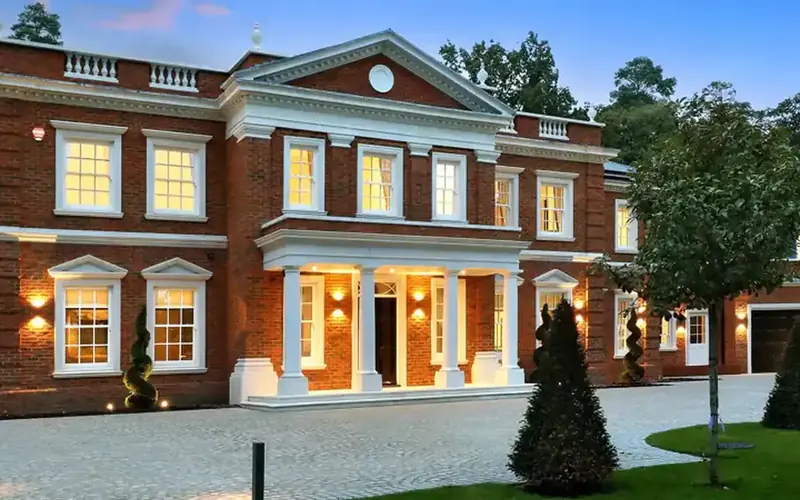 Large brick house with a columned porch, a block paved drive and warm light in the windows